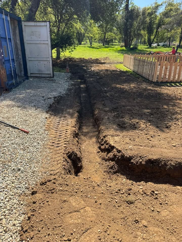 A long, narrow dirt trench dug into the ground next to a gravel path, a shipping container, and a wooden fence.