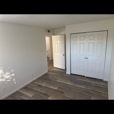 Empty bedroom with wood-look flooring, white walls, closet doors, and an open doorway to a bathroom