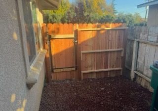 Narrow backyard with wooden fence and gate beside a house, covered in dark gravel.