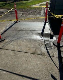 Sidewalk blocked by red posts and yellow caution tape around a damaged concrete slab.
