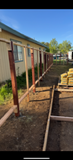 Side view of a mobile home with red posts beside a dirt path and stacked lumber outdoors