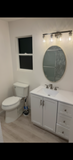Small bathroom with white vanity, oval mirror, toilet, window, and light wood floor.