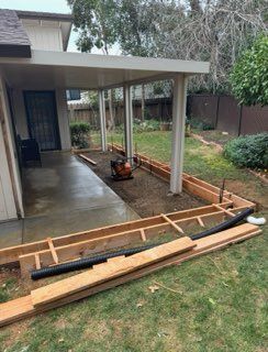 Backyard patio under construction with wooden framing, concrete slab, and stacked boards beside a house