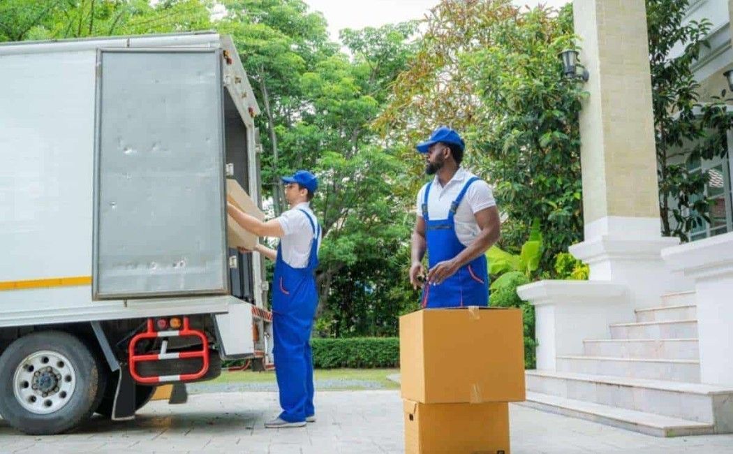 Two movers in blue uniforms loading a truck beside a house, with moving boxes nearby