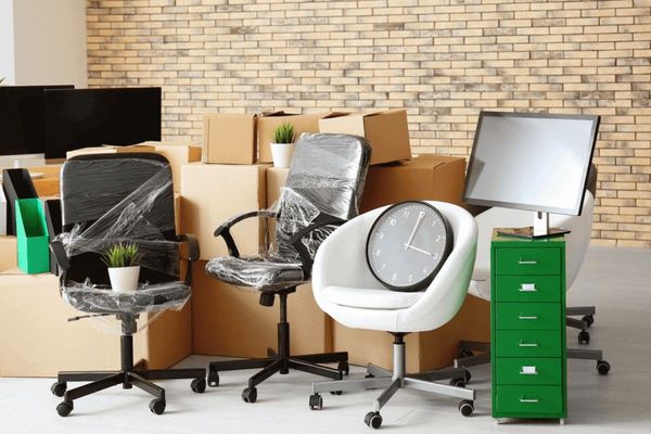 Office chairs and desks arranged in a storage area, with boxes and a green filing cabinet against a brick wall. Office chairs and desks arranged in a storage area, with boxes and a green filing cabinet against a brick wall.
