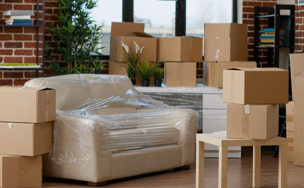 Living room with wrapped sofa and stacked moving boxes, suggesting a home in transition