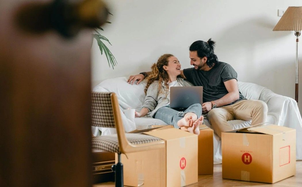 Couple sitting on a couch among moving boxes, smiling at a laptop in a bright living room. Couple sitting on a couch among moving boxes, smiling at a laptop in a bright living room.