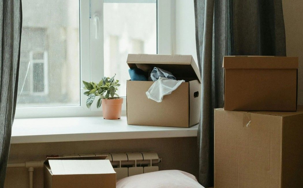 Boxes by a window in a room, with a small plant and curtains nearby. Boxes by a window in a room, with a small plant and curtains nearby.