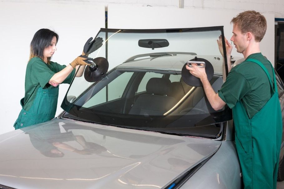 Two people installing a windshield on a silver car in a garage