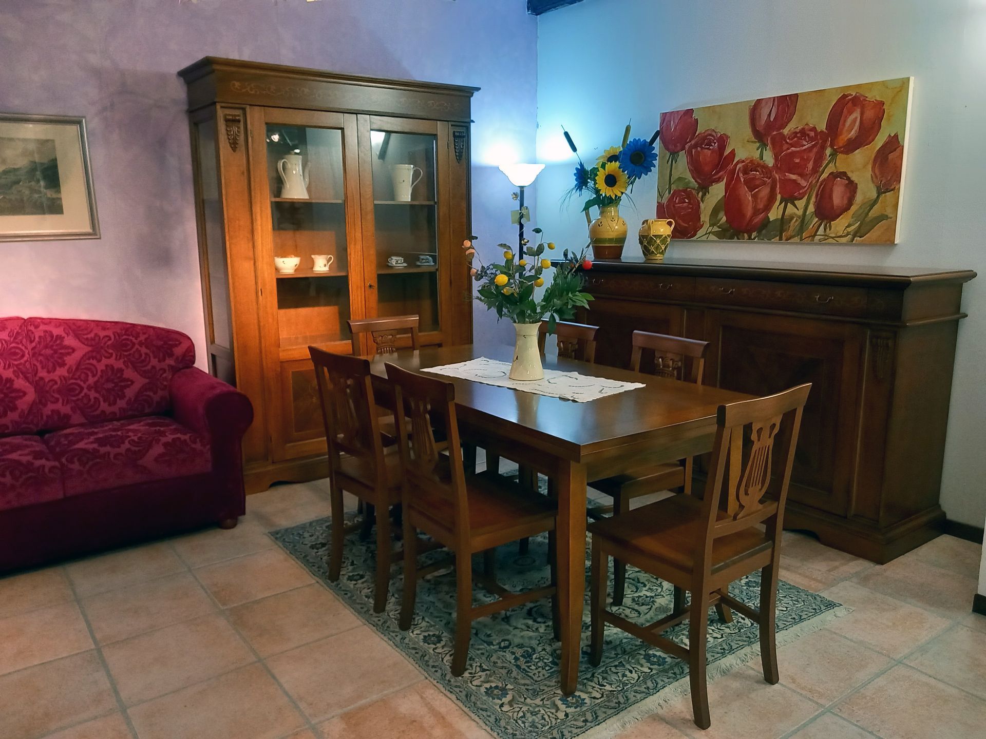 Sala da pranzo con mobili in legno, tra cui tavolo, sedie, credenza e credenza. Sono presenti anche un divano rosso e opere d'arte floreali.