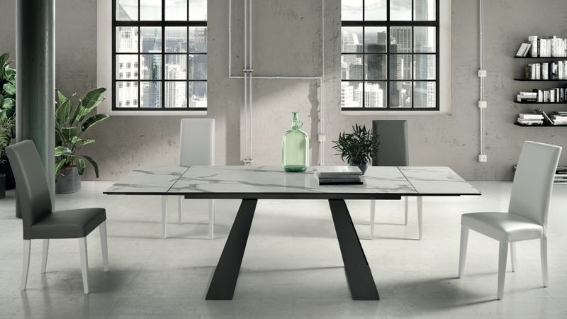Sala da pranzo moderna con tavolo con piano in marmo, base scura e quattro sedie bianche. Ampie finestre e ambiente industriale.