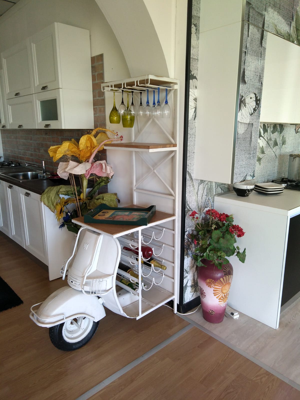 Portabottiglie bianco a forma di scooter in cucina, con bottiglie, bicchieri e oggetti decorativi.