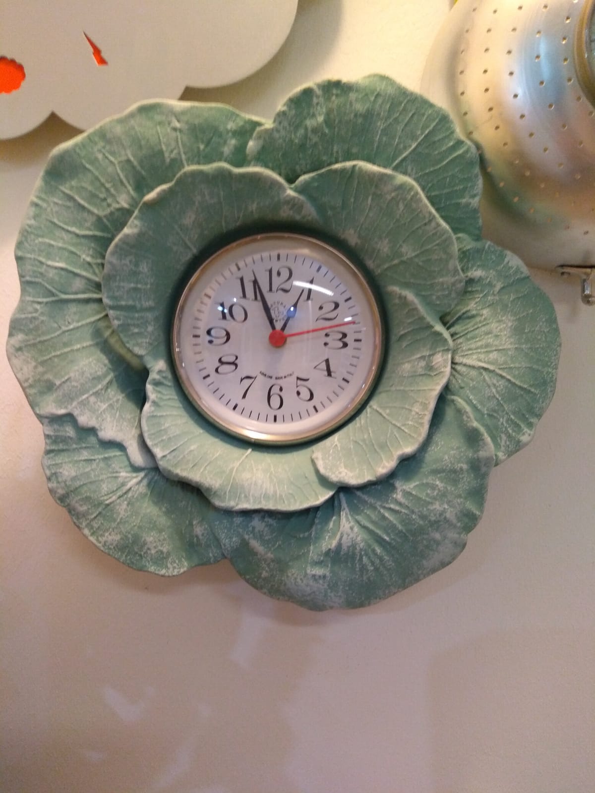 Un orologio da parete a forma di fiore di cavolo; il quadrante è bianco e il fiore è verde chiaro.