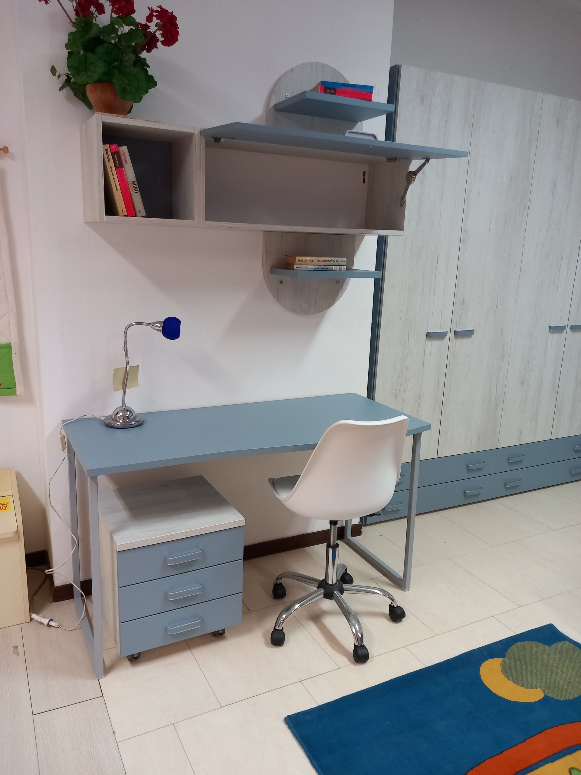 Una scrivania grigia, un mobile contenitore e degli scaffali fissati a una parete con una sedia bianca e un tappeto.