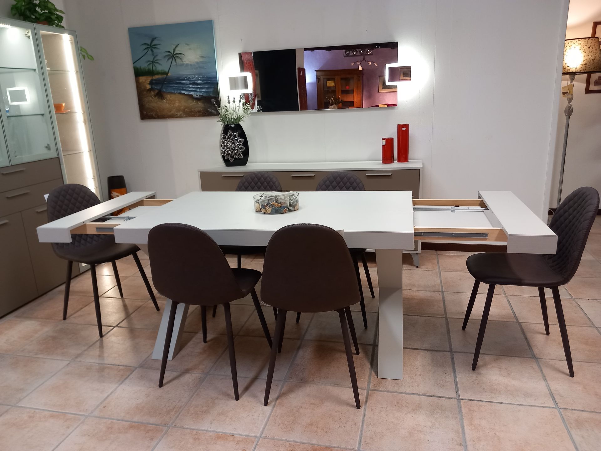 Sala da pranzo con tavolo bianco allungabile, sedie marroni, credenza e specchio.