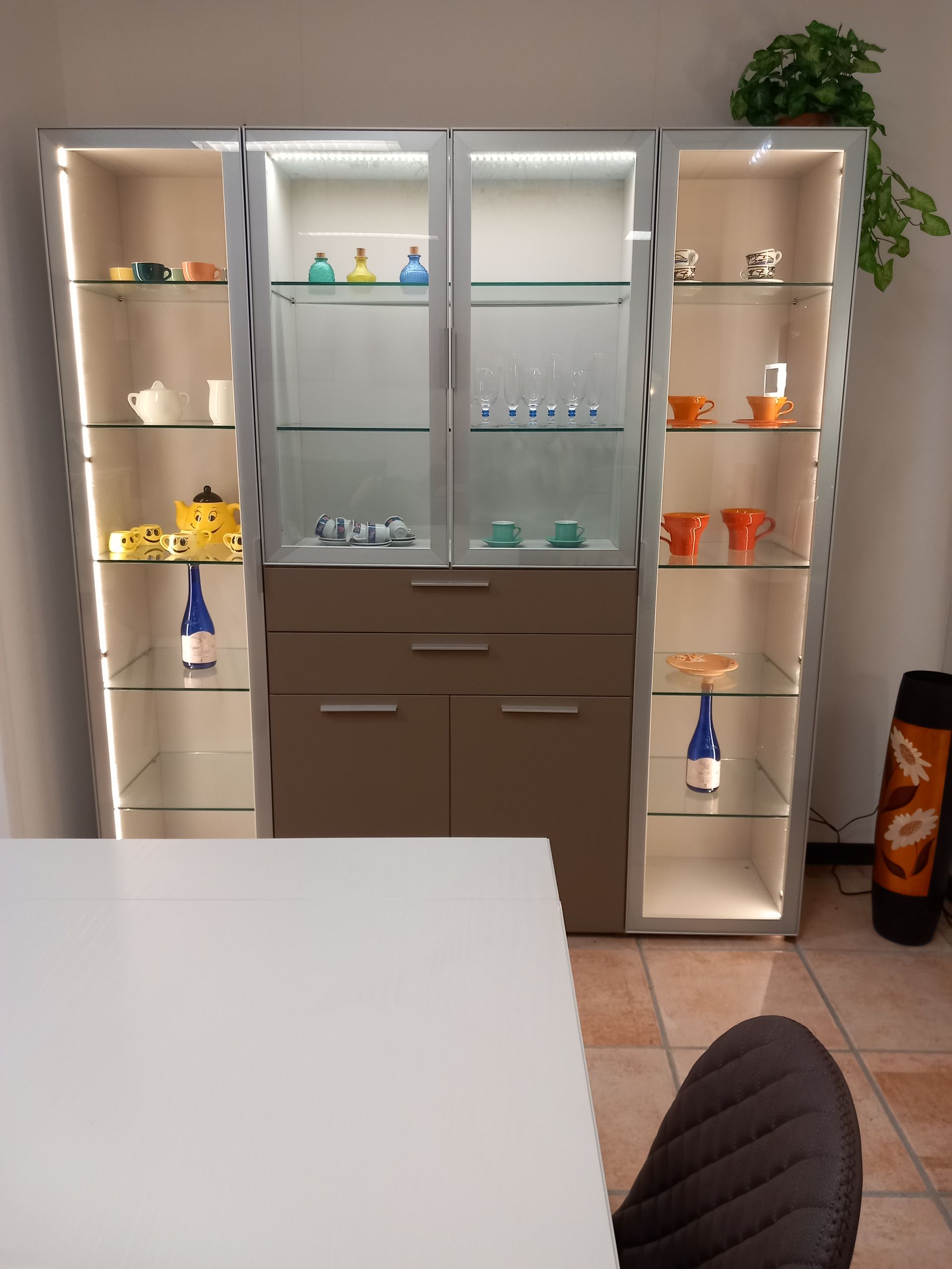 Vetrina con ripiani in vetro, interno illuminato, contenente tazze e piccoli oggetti, appoggiata a una parete.
