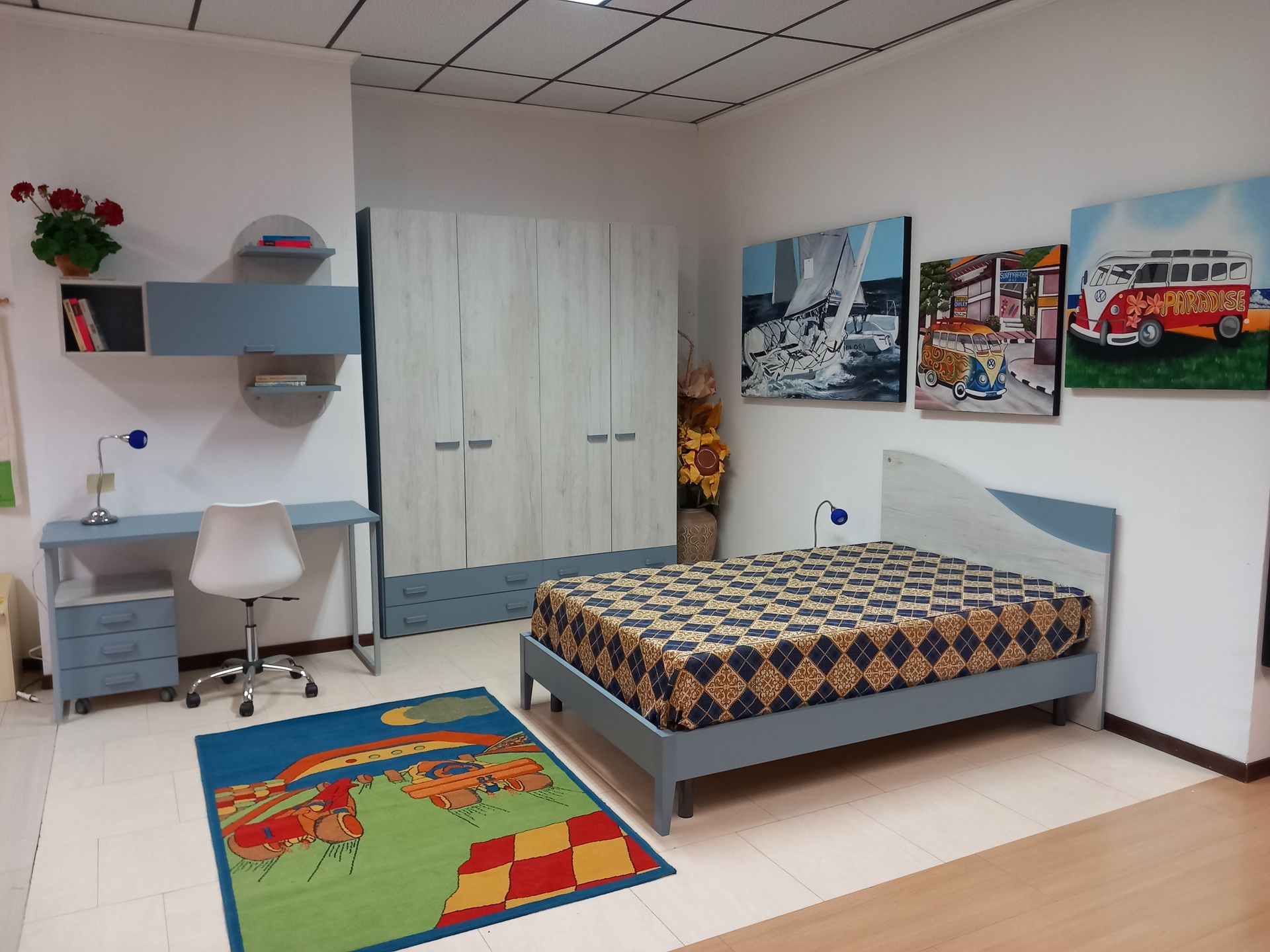 Camera da letto con mobili blu e bianchi, letto, scrivania, armadio, quadri e tappeto colorato.