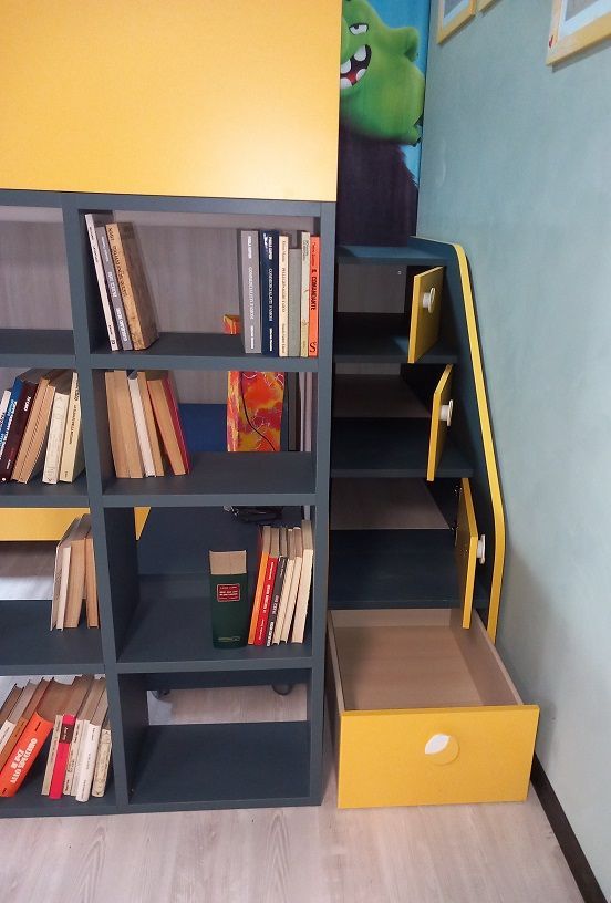 Libreria integrata con ripiani a cubo e gradini che conducono alla zona living. Dettagli gialli. Libri e un cassetto portaoggetti sono visibili.