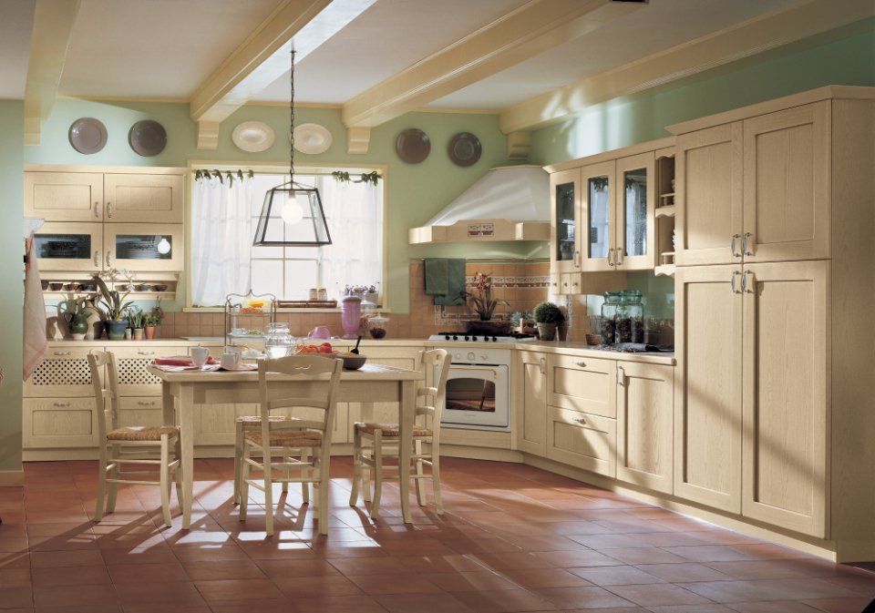 Cucina beige con pareti verdi, travi in legno e pavimento in cotto. Include mobili, tavolo ed elettrodomestici.