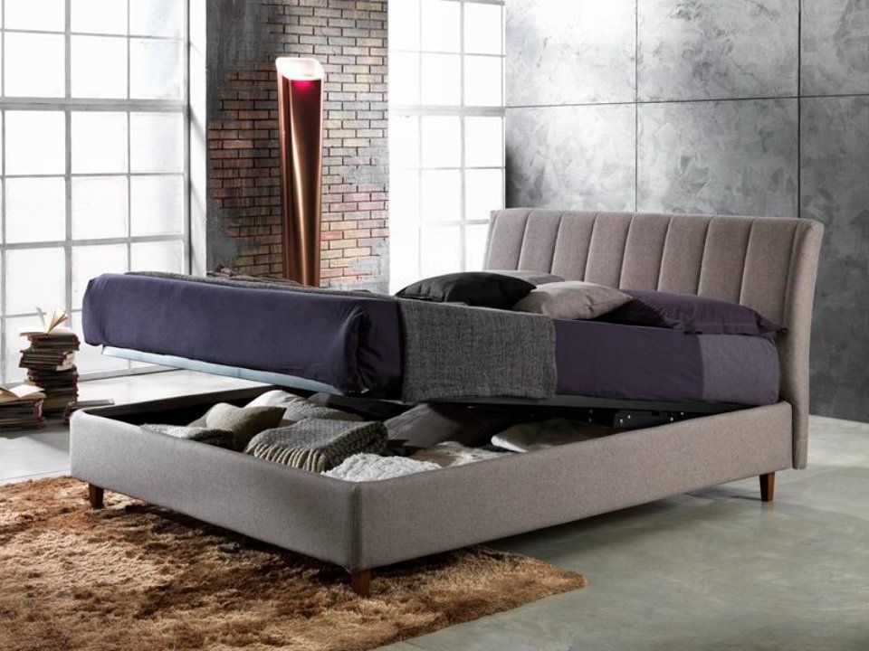 Struttura del letto imbottita con materasso rialzato che rivela un vano contenitore; colori neutri, ambiente industriale.