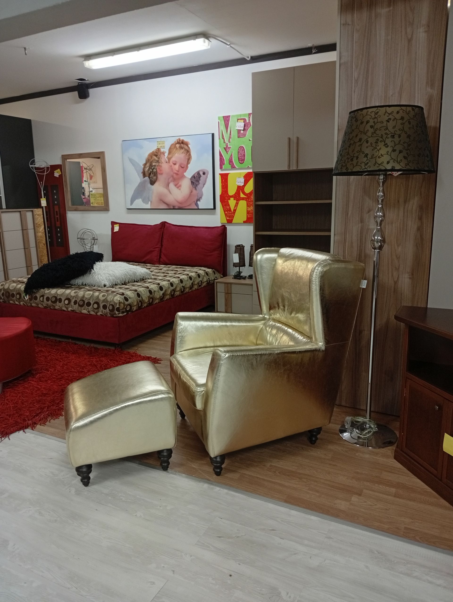 Poltrona e pouf dorati in uno showroom di mobili con letto rosso, opere d'arte e lampada da terra.