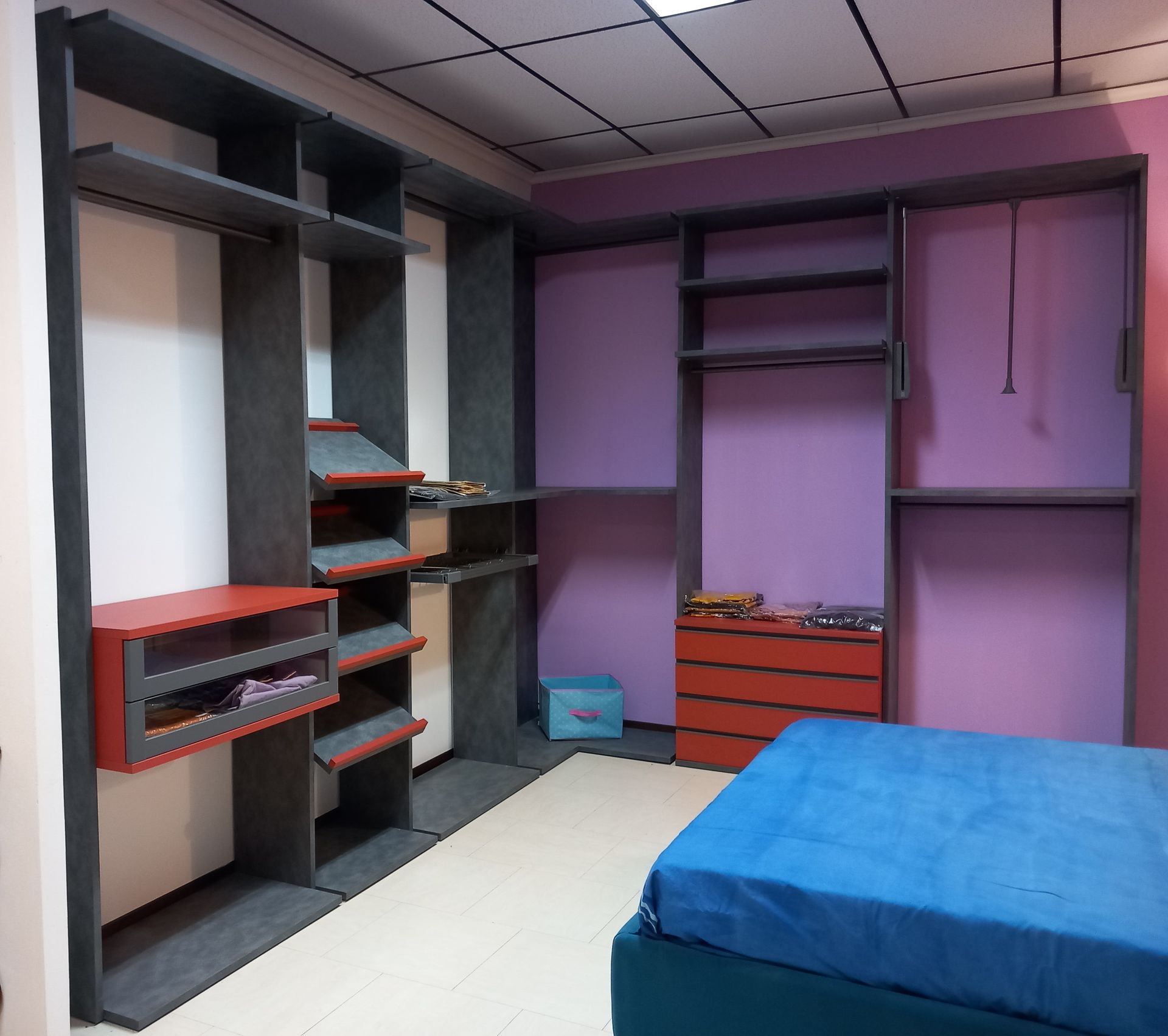 Cabina armadio vuota con scaffali grigi e rossi e un letto blu. Pareti viola e moquette color crema.