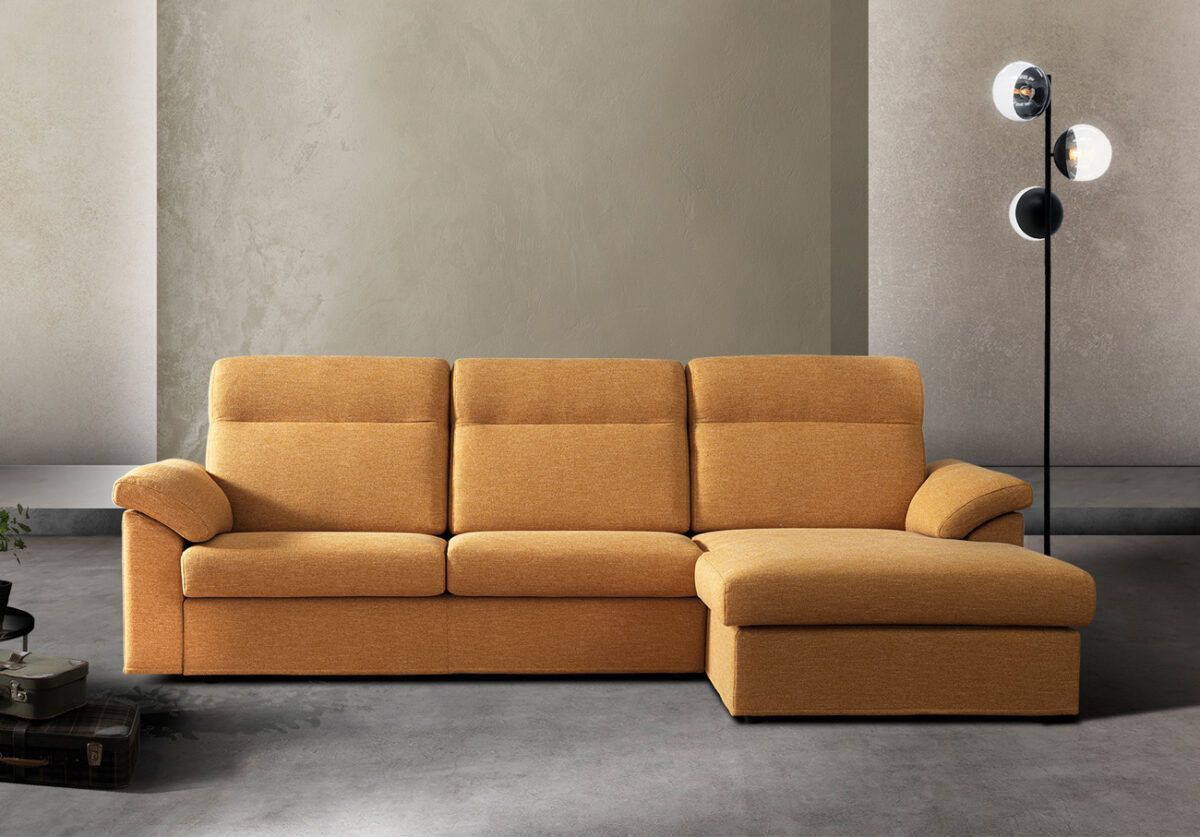 Divano componibile giallo con chaise longue contro una parete color cuoio, accanto a una lampada da terra nera.