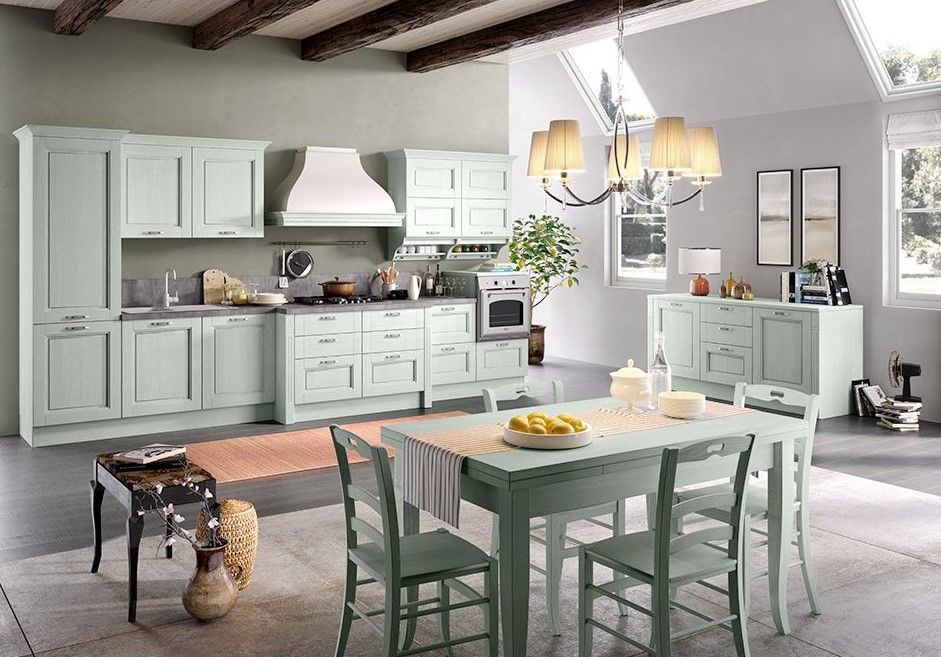 Cucina verde chiaro con mobili, tavolo e sedie. Cappa, lampadario e finestre bianchi.