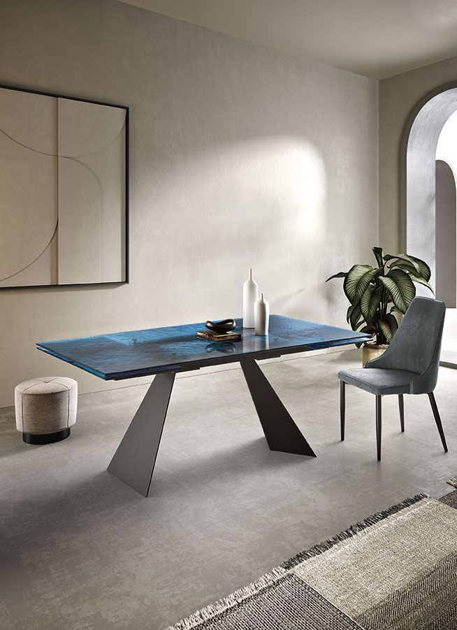 Tavolo da pranzo con piano blu e gambe angolate grigie, in una moderna sala da pranzo con sedia, pianta e opere d'arte.