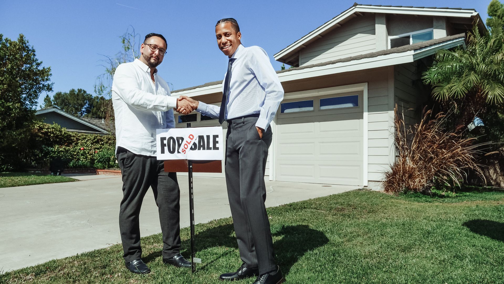 Two men shake hands beside a “For Sale” sign in front of a suburban house and garage.