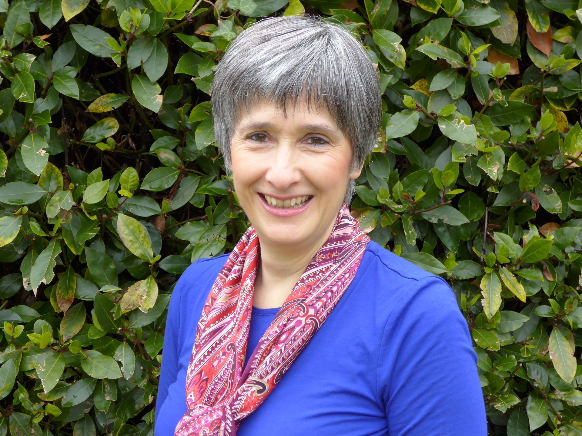 A woman in a brown jacket and necklace is standing in front of a plant.