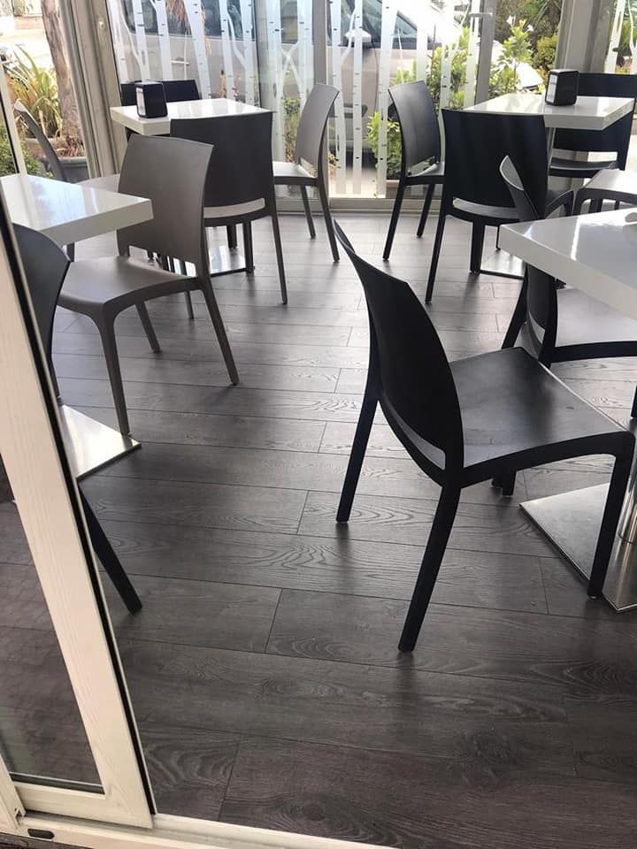 Interno di un bar con tavoli bianchi e sedie nere e grigie su pavimento in legno scuro.