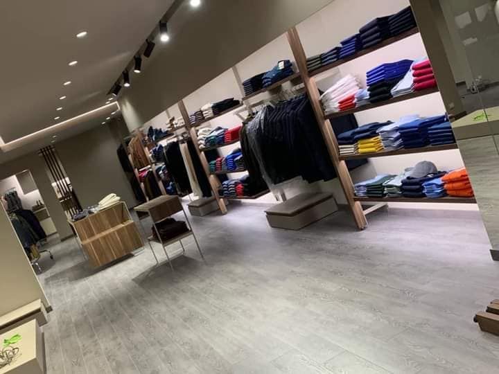 Interno di un negozio di abbigliamento con appendiabiti, scaffali con vestiti piegati e pavimento in legno chiaro.