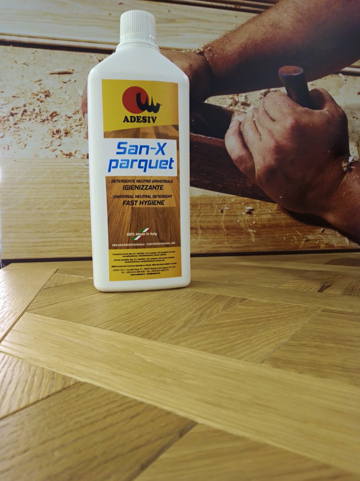 Flacone di soluzione detergente per parquet San-X su pavimento in parquet, con un falegname che lavora sul legno sullo sfondo.