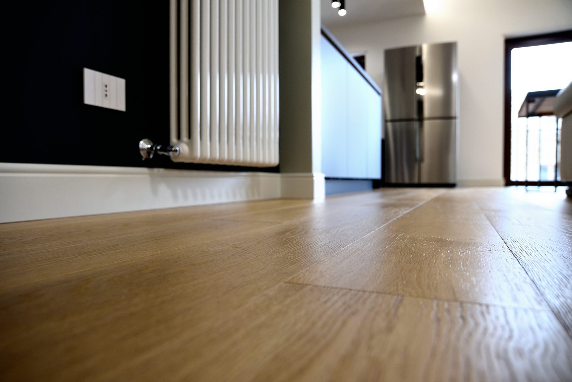 Vista dal basso di un pavimento in legno chiaro in un interno moderno, con termosifone, parete nera ed elettrodomestici sullo sfondo.