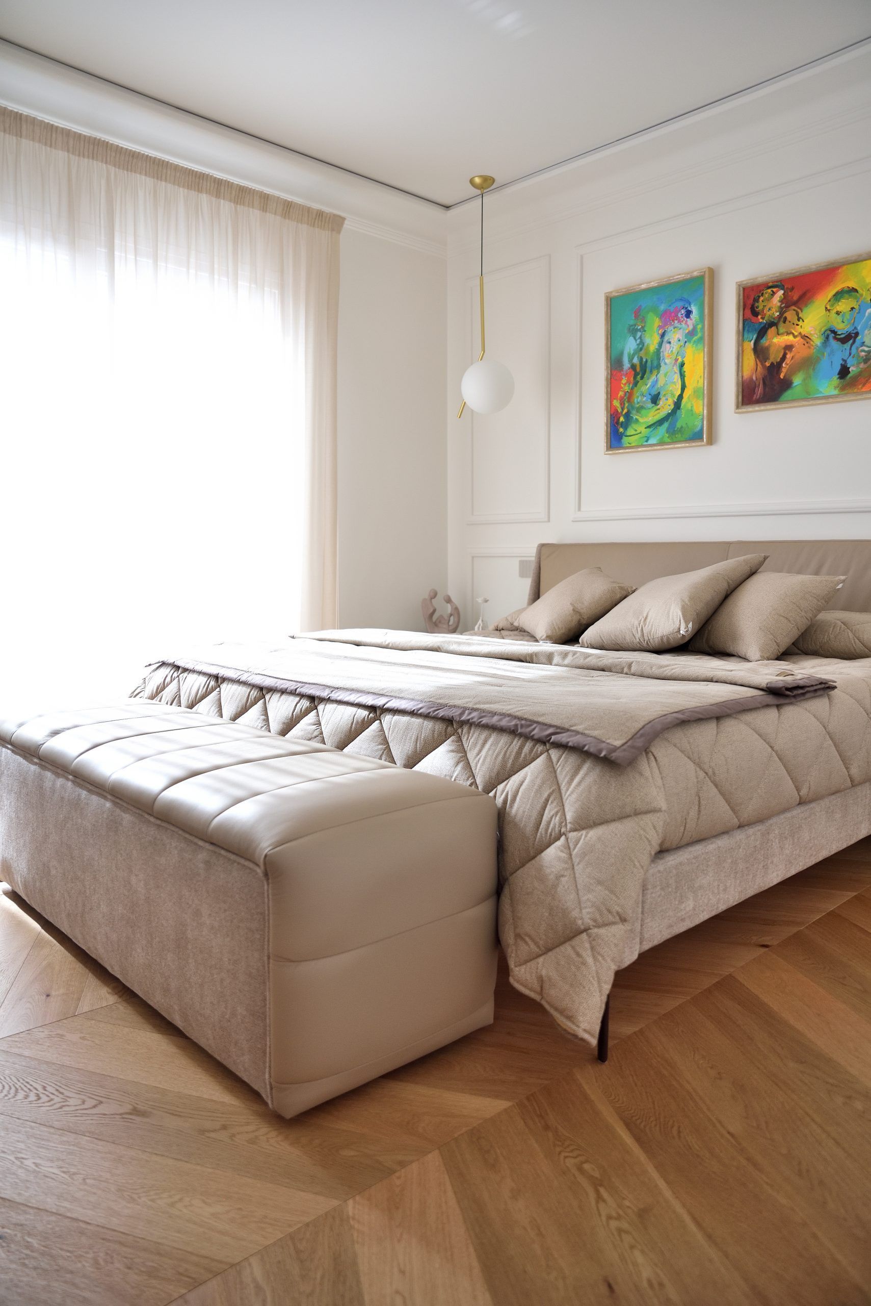 Elegante camera da letto con un letto grande, biancheria beige e una panca trapuntata ai piedi. Due quadri colorati alle pareti.