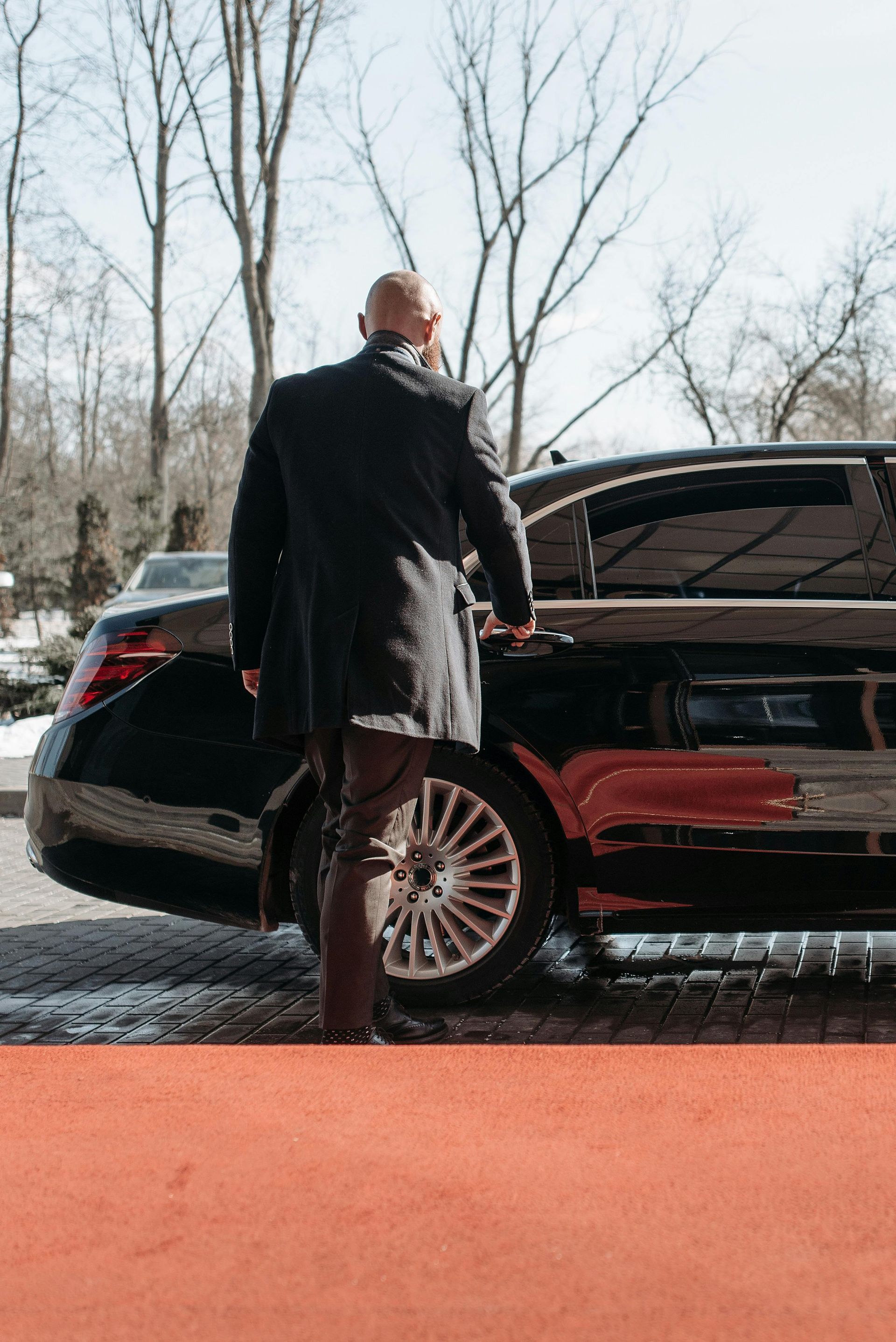 Man in a dark coat exiting a black car on a red carpet. Sunlight, outdoor setting.