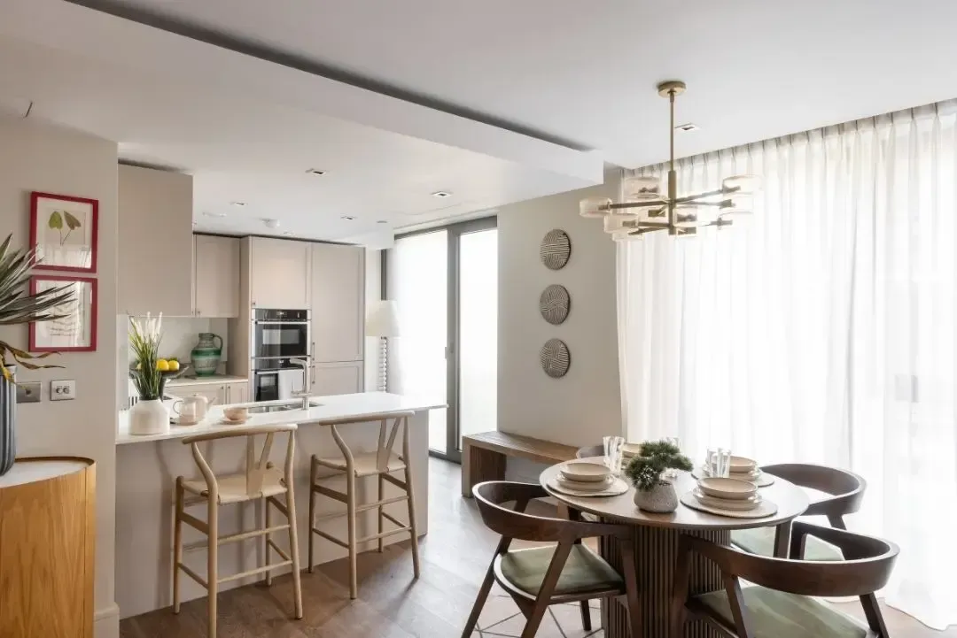 Bright open kitchen and dining area with white walls, bar stools, and a round table by large windows.