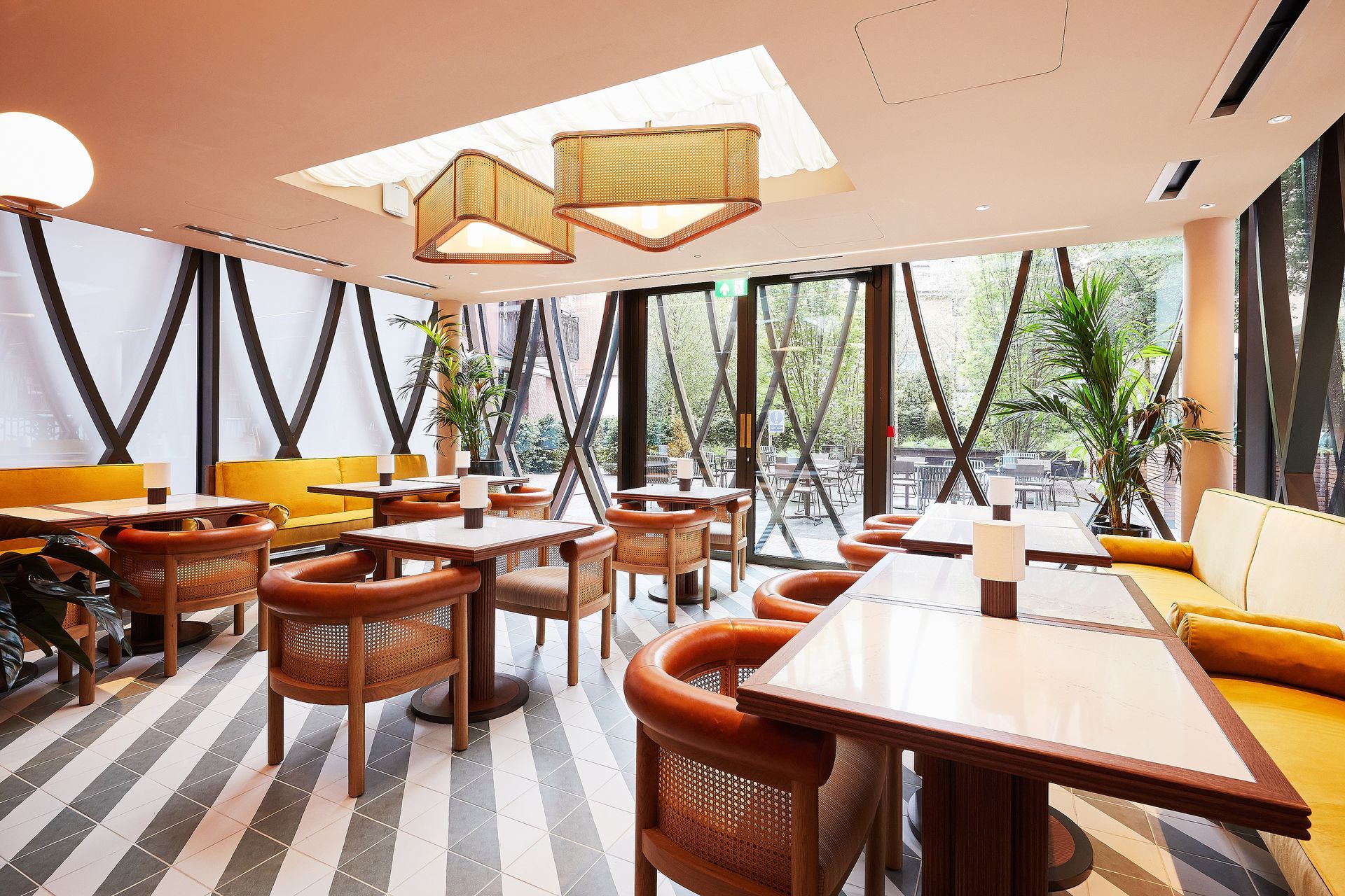 Modern restaurant interior with wooden tables, woven chairs, large windows, and geometric pendant lights.