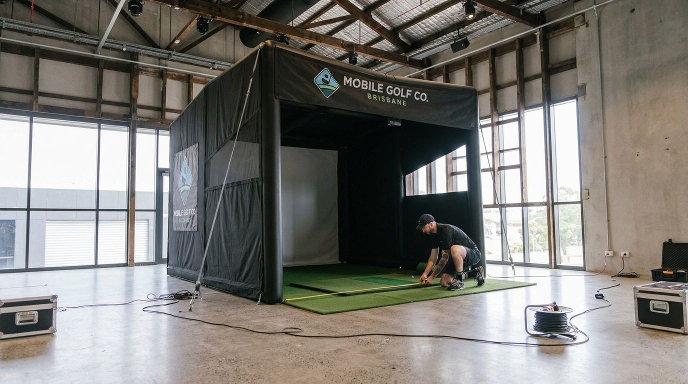Worker setting up a black golf simulator booth with green turf inside a warehouse venue