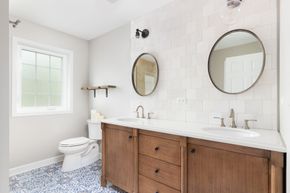 A bathroom with a double wood vanity, white tile backsplash, two circular mirrors, and patterned blue and white floor tile.