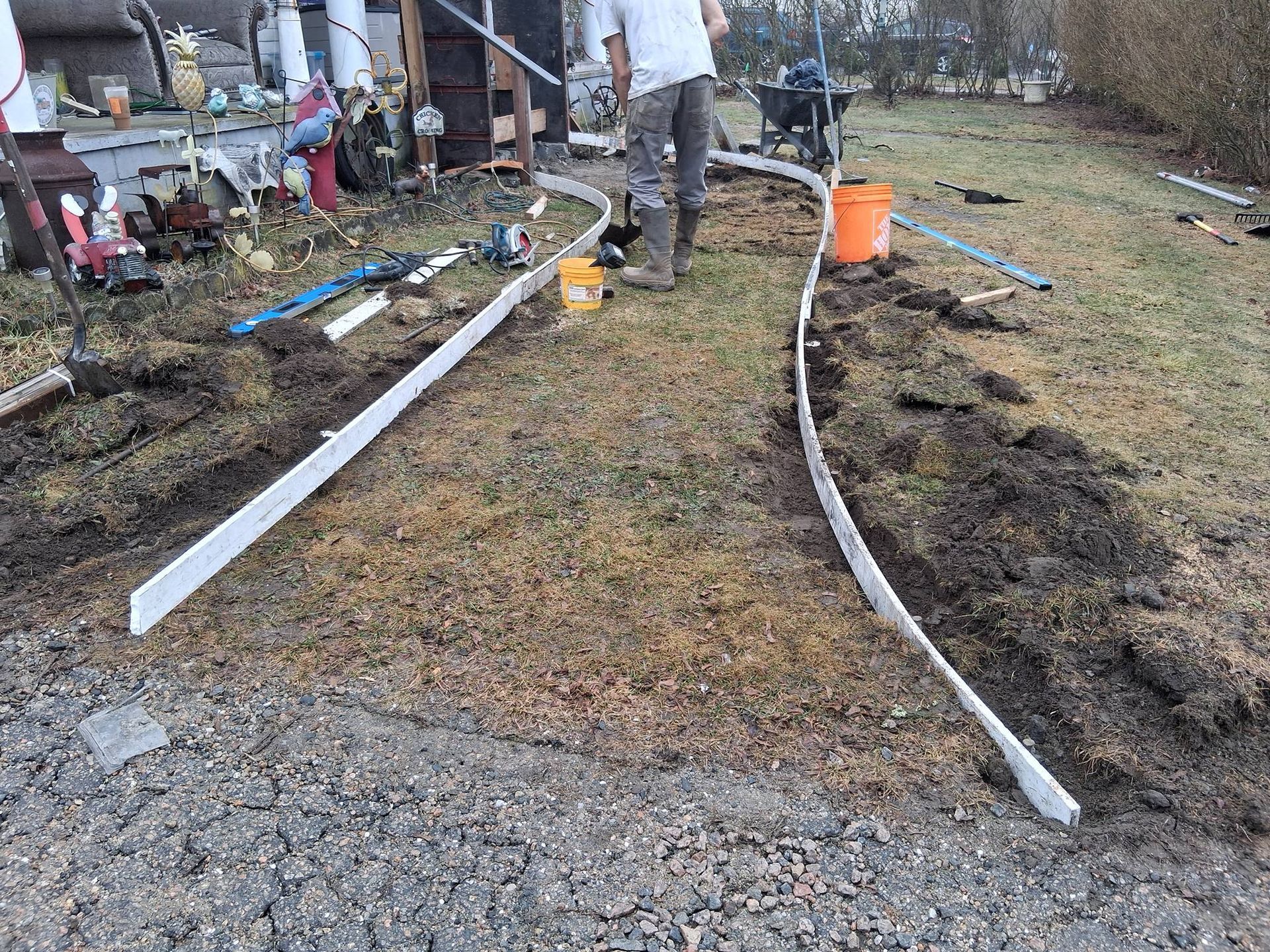 A worker installs white landscape edging along a path in a yard with gravel and turned soil.