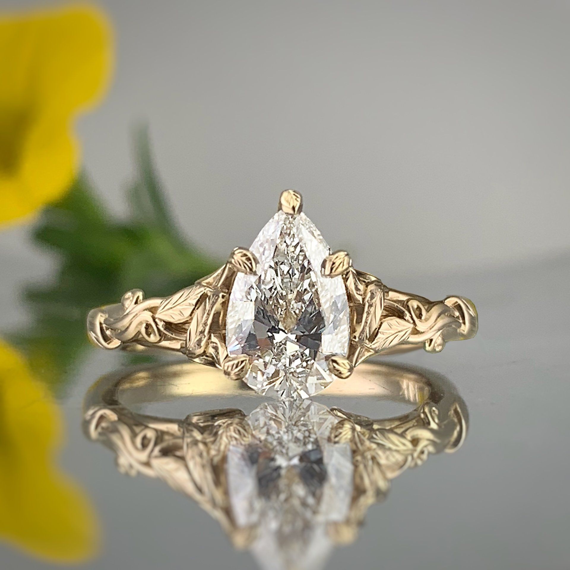A pear shaped diamond ring is sitting on a table next to a yellow flower.