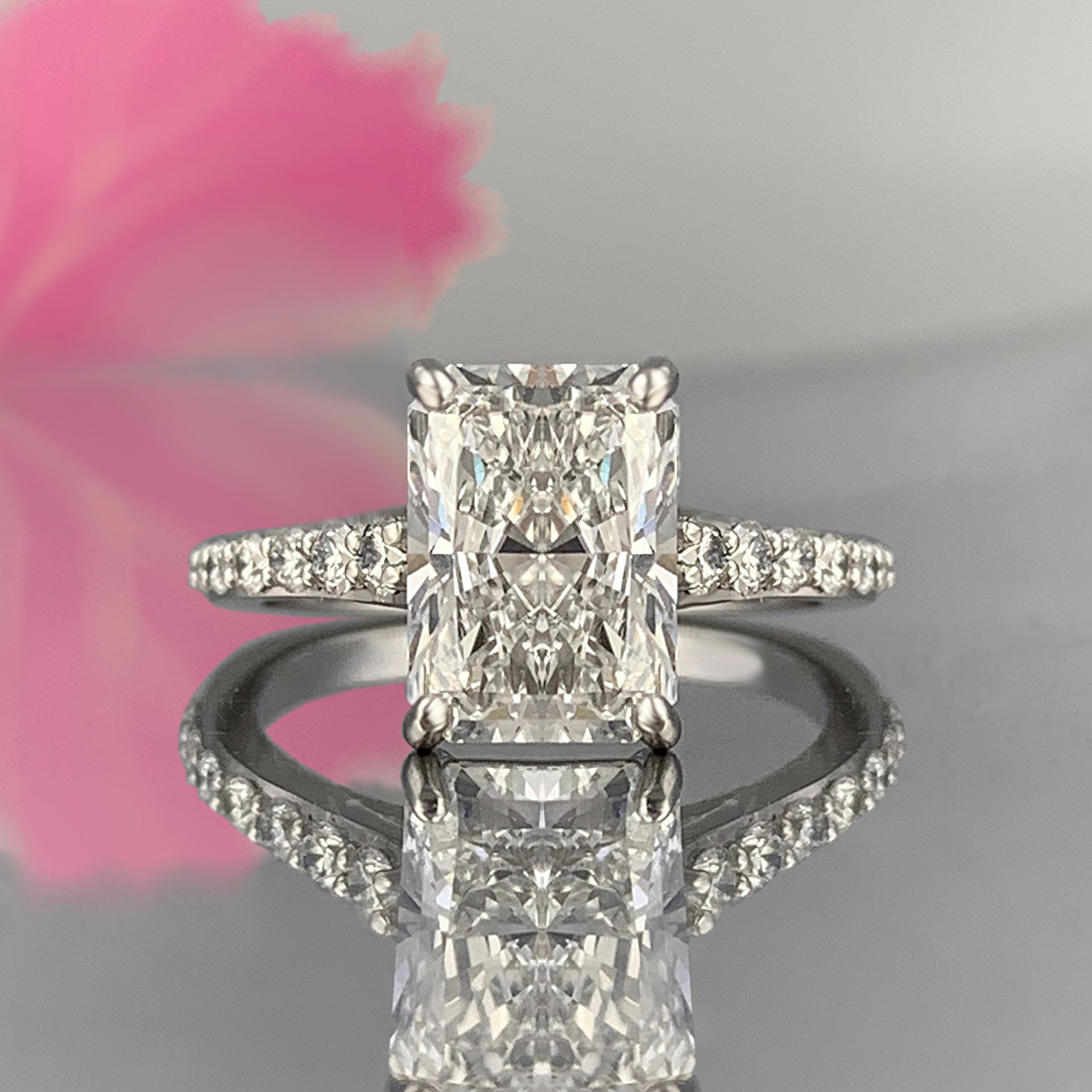 A diamond ring is sitting on a table with a pink flower in the background.