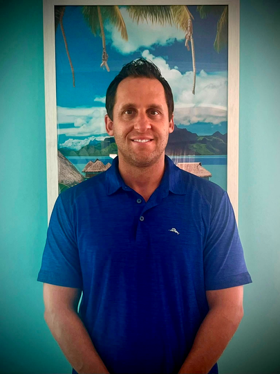 Man in a blue polo shirt smiling in front of a tropical beach painting
