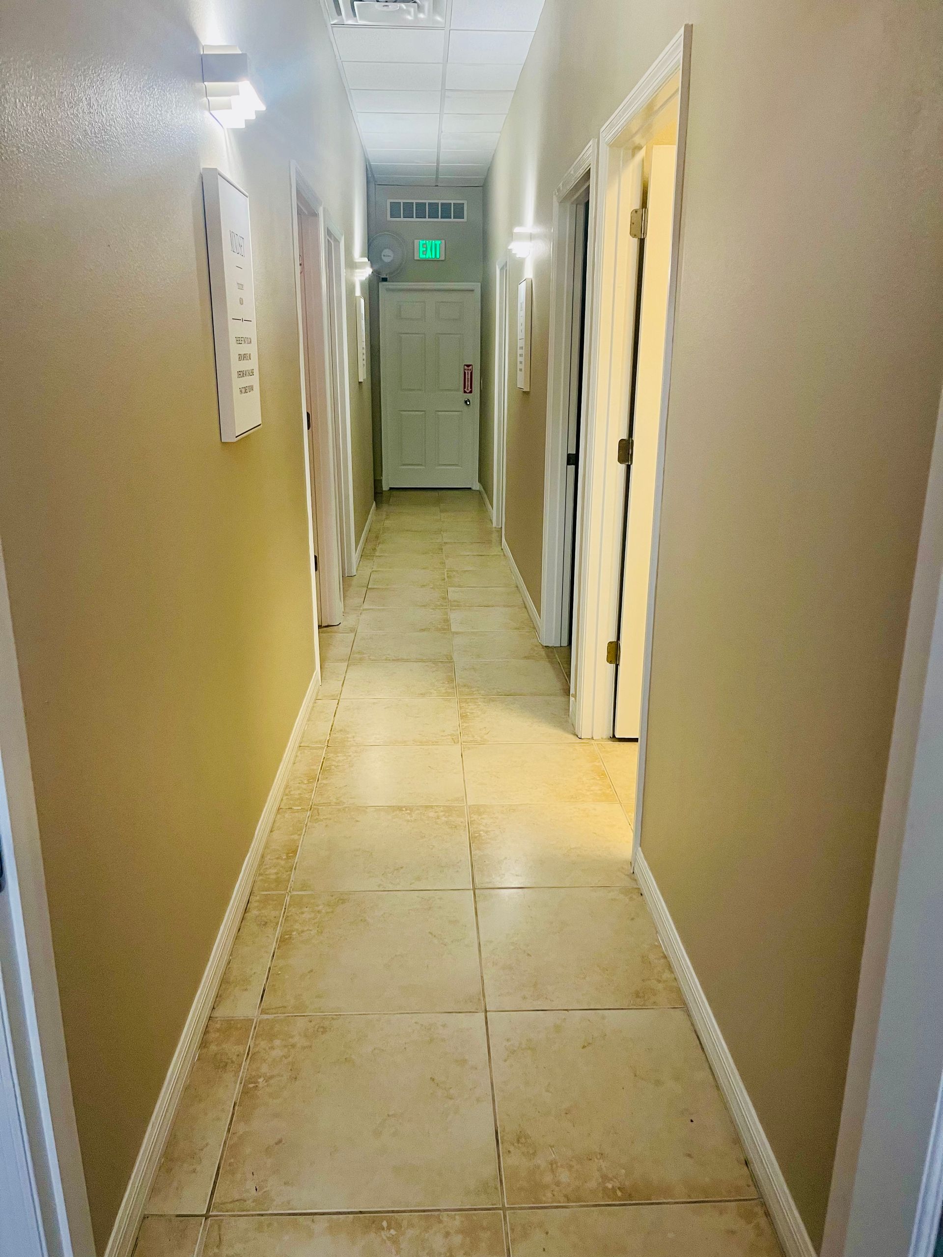 Narrow beige hallway with tiled floor and doors along both sides, ending at a closed door with exit sign.
