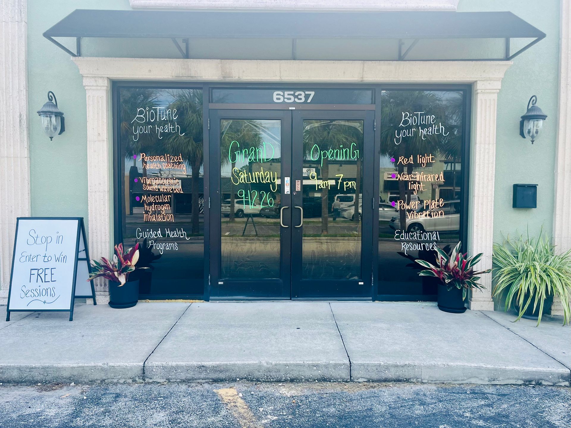 Storefront entrance with double glass doors, potted plants, and a sidewalk sign under an awning