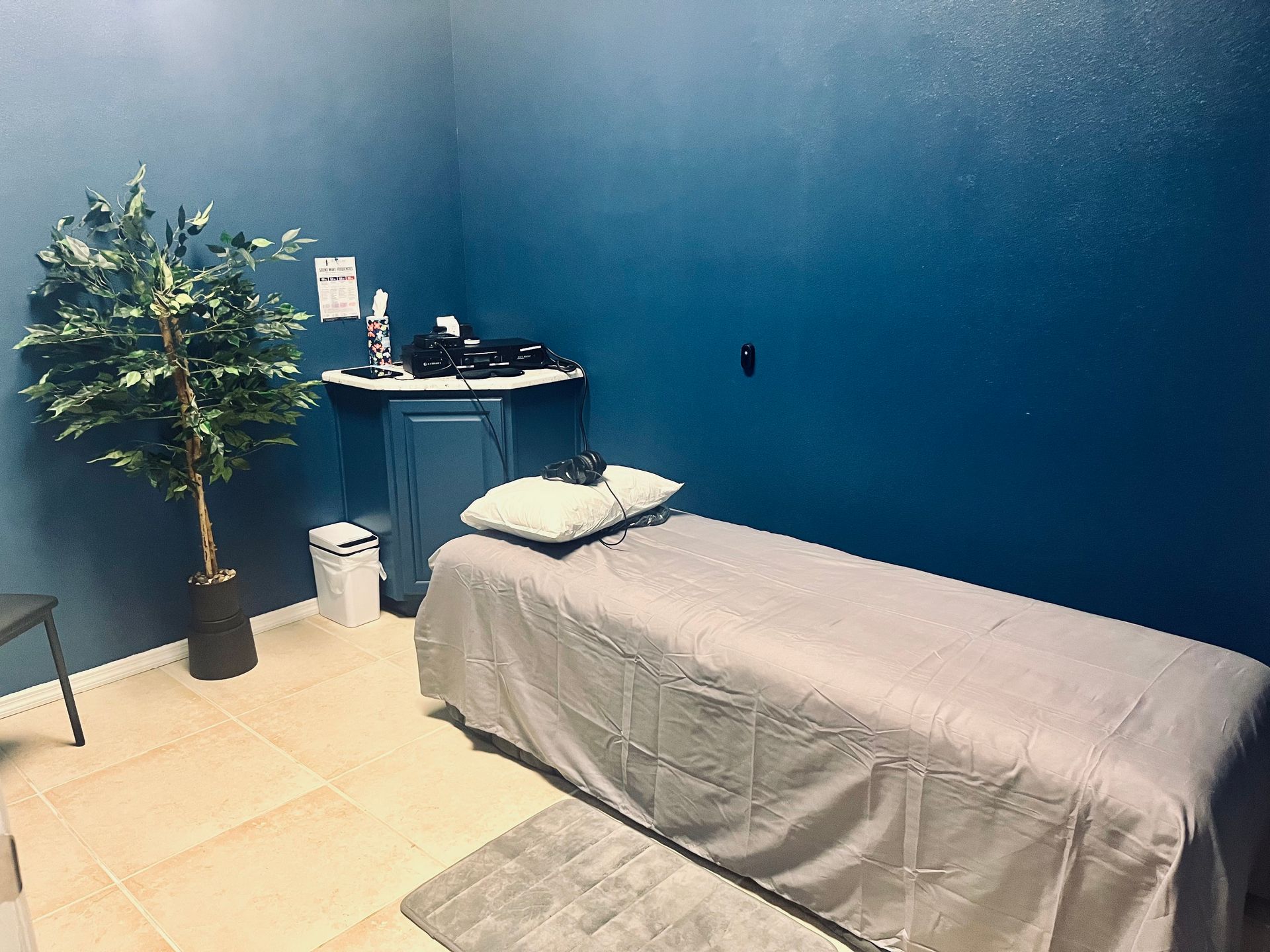 Massage room with blue walls, treatment table, small desk, and a potted plant.