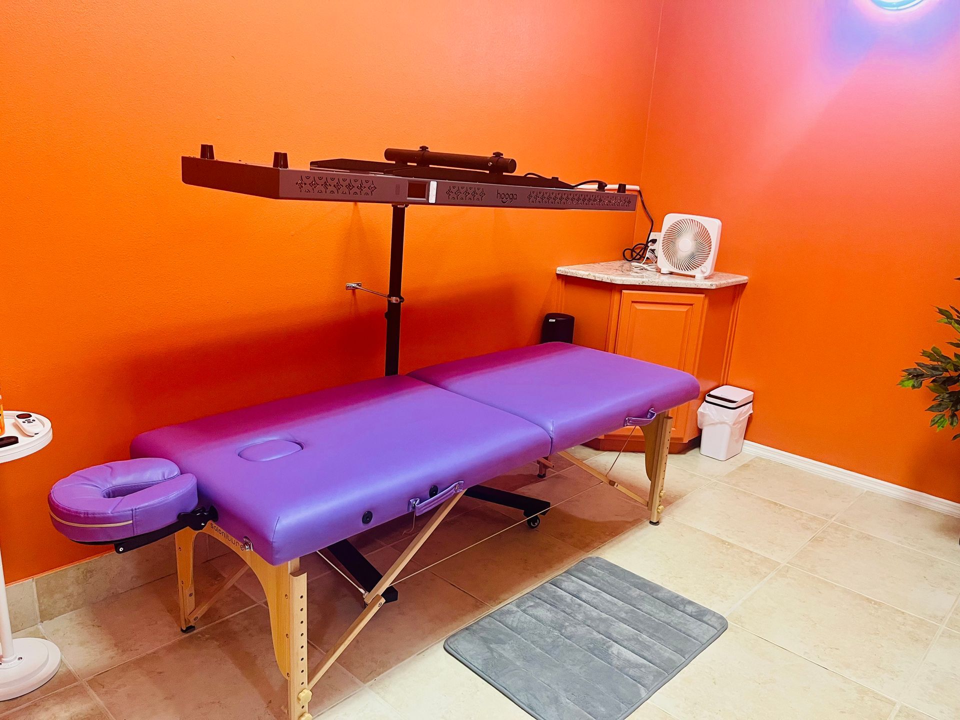 Massage table in a bright orange treatment room with a black shelf and gray rug.