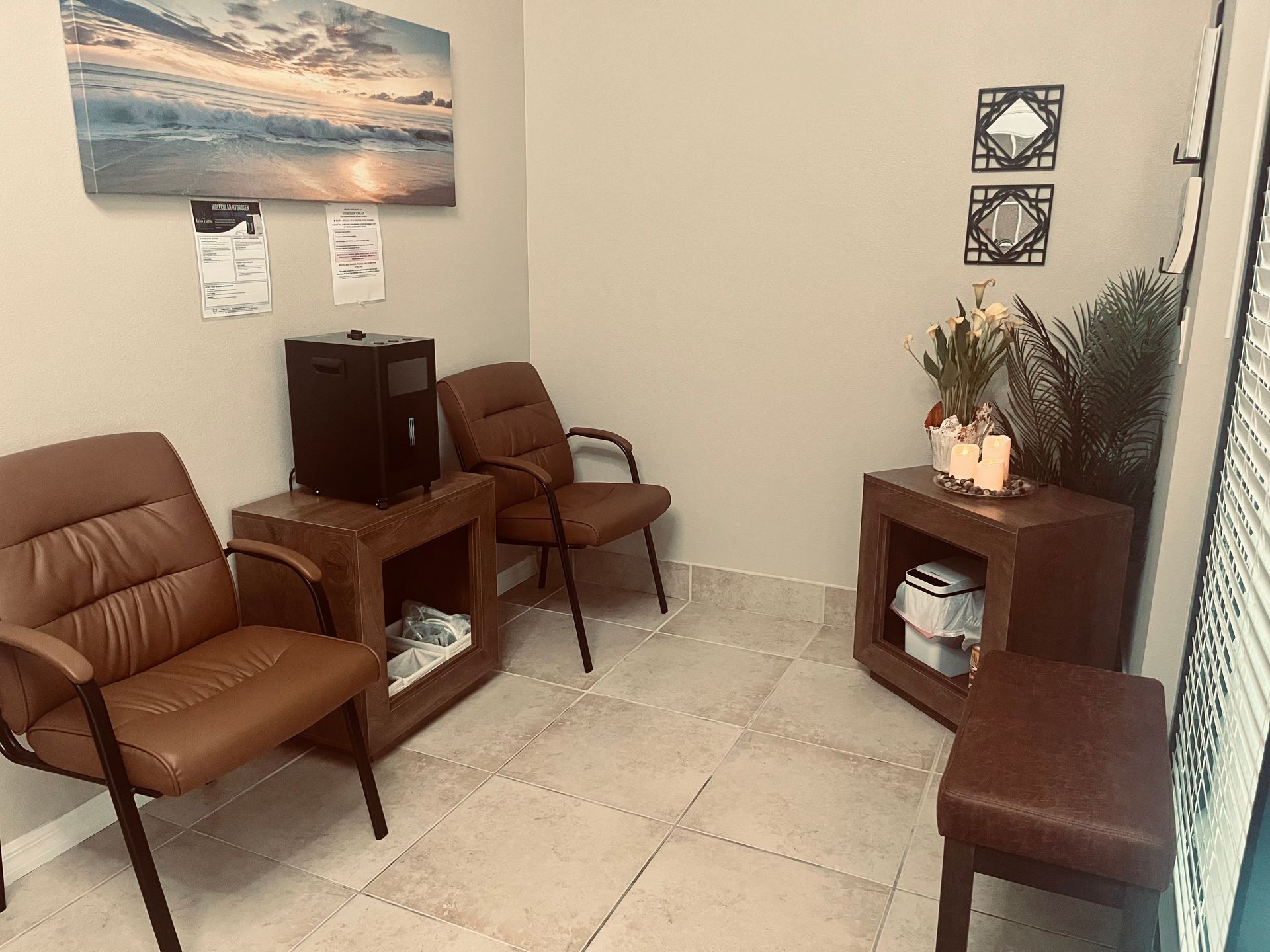 Small waiting room with brown chairs, side tables, a wall picture, and a potted plant on tile floor.